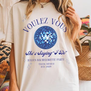 May include: Cream-colored t-shirt with the text "VOULEZ VOUS" above a blue disco ball graphic. Below the graphic, the text reads "She's Saying 'I Do'" and details for Julia's bachelorette party in Tulum, Mexico, July 2029. The shirt has short sleeves.