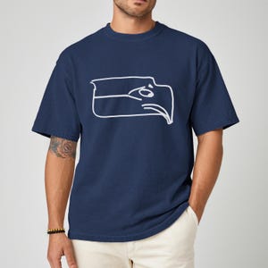 Puede incluir: Camiseta azul marino con un contorno blanco de la cabeza de un &aacute;guila. La camiseta de manga corta tiene cuello redondo y un corte relajado. El gr&aacute;fico es un dise&ntilde;o simple y moderno. La camiseta se combina con pantalones de color claro.