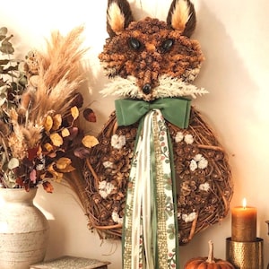 May include: A fox-shaped wall hanging crafted from brown pinecones and twigs, featuring a green fabric bow with long, trailing ribbons. The fox head has dark eyes and white accents, while the body is a circular wreath adorned with small white flowers.