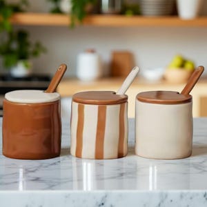Puede incluir: Tres botes de cerámica para especias con tapas y cucharas. Uno es marrón liso, otro tiene rayas marrones y el tercero es crema liso. Las tapas son marrones y las cucharas son crema y marrón. Los botes están sobre una encimera de mármol blanco.