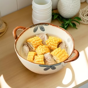 以下が含まれることがあります： トウモロコシと肉片が入ったスープが入ったセラミックボウル。ボウルには2つのハンドルがあり、花柄で装飾されています。ボウルは木製の表面に置かれています。