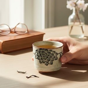 May include: A ceramic teacup with a black floral design, filled with tea, is held by a person. The cup is off-white with a brown base. A book and glasses are in the background. The tea cup is a beautiful piece of pottery.