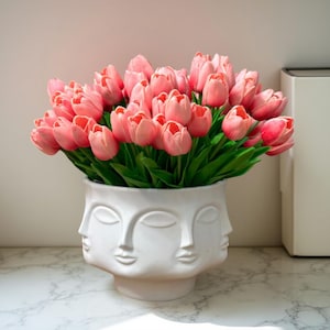 Pode incluir: Um vaso de cerâmica branco com múltiplos rostos contém um grande buquê de tulipas rosa com folhas verdes. O vaso está sobre uma superfície com padrão de mármore. As tulipas estão em plena floração, criando um arranjo floral vibrante.