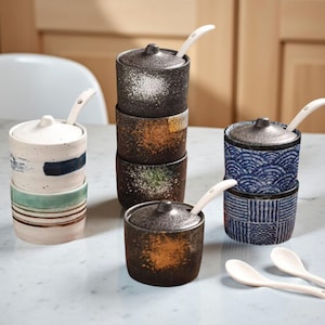 Könnte beinhalten: Eine Sammlung von Keramik-Gewürzgläsern mit Deckeln und kleinen Löffeln. Die Gläser sind in verschiedenen Farben und Mustern gehalten, darunter gesprenkeltes Schwarz, Orange, Grün und Blau mit weißen Akzenten. Die Löffel sind weiß.