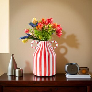 May include: A red and white striped ceramic vase with two handles, filled with a colorful bouquet of flowers. The vase is on a dark wooden surface, with a silver bottle and small metal cylinder to the left.