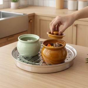 May include: Two ceramic cooking pots, one light green and one brown, sit on a metal tray. The brown pot contains food and is being covered with a lid. The pots are on a wooden surface, suggesting a kitchen setting.