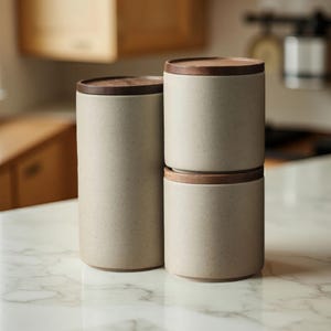 Puede incluir: Un juego de tres botes de cerámica beige con tapas de madera marrón oscuro. Los botes cilíndricos tienen una textura moteada. Uno está solo, los otros dos están apilados. Los recipientes de cocina están sobre una encimera de mármol.