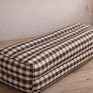 May include: A rectangular brown and white checkered yoga bolster pillow. The pillow is made of a soft fabric and is designed for comfort and support during yoga practice. The bolster is placed on a wooden surface.