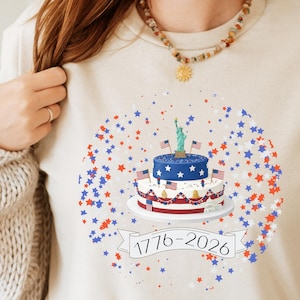 May include: Beige t-shirt featuring a patriotic cake design with American flags and the Statue of Liberty. The cake is surrounded by red, white, and blue stars. The text "1776-2026" is displayed on a banner below the cake.