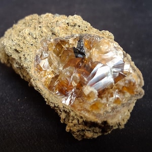 May include: A raw mineral specimen featuring a rough, textured, tan-colored exterior crust that encases a cluster of translucent, amber-colored crystals. The crystalline center displays multifaceted surfaces that reflect light with a glossy finish.