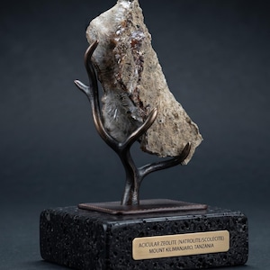 May include: A specimen of acicular zeolite, specifically natrolite and scolecite from Mount Kilimanjaro, Tanzania, mounted on a dark bronze-colored branch-shaped stand atop a rectangular black volcanic rock base with a gold-toned identification plaque.