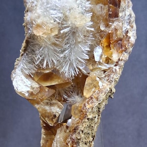 May include: A mineral specimen featuring a cluster of translucent amber-colored crystals and white, needle-like radiating crystal formations embedded in a rough, textured, earthy-brown rock matrix, displayed on a clear plastic stand.