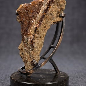 May include: A raw, amber-colored mineral specimen with crystalline textures mounted on a curved, dark bronze-toned metal stand. The stand is secured to a circular, polished dark brown stone base featuring subtle fossilized inclusions.