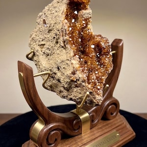 May include: An amber crystal geode with a rough, textured exterior and sparkling orange-brown interior crystals. The geode is mounted on a dark wood stand with gold-toned metal supports and a plaque that reads AMBER CRYSTAL GEODE.