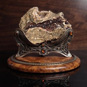 May include: A rough, textured mineral specimen with dark brown crystalline inclusions, cradled in an ornate, antique-finished metal stand featuring amber-colored cabochons, all mounted on a polished, circular wooden base with a decorative metal rim.