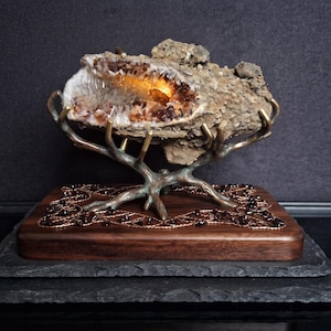 May include: A raw citrine geode with a glowing orange interior, resting on a bronze-colored metal stand shaped like branches. The stand is mounted on a rectangular wooden base featuring a decorative copper wire inlay with small dark beads.