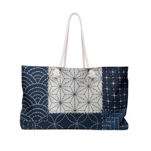 May include: A large tote bag featuring a patchwork design of dark blue and white fabric with various geometric patterns including waves, stars, and circles. The bag has two white rope handles attached with metal grommets at the top edge.
