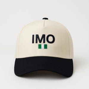 Op de afbeelding: Crèmekleurige baseballcap met een zwarte klep. De cap heeft het woord "IMO" in zwarte blokletters, met daaronder een klein groen-wit vlagontwerp.