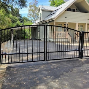 May include: Black metal driveway gate with a matching pedestrian gate. The gates have a curved top design and vertical bars. The gates are in front of a house with a light-colored exterior and a paved driveway.