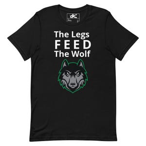 Puede incluir: Camiseta negra con el texto blanco "The Legs FEED The Wolf". Debajo, un gráfico de cabeza de lobo gris y verde. La camiseta tiene cuello redondo y mangas cortas.
