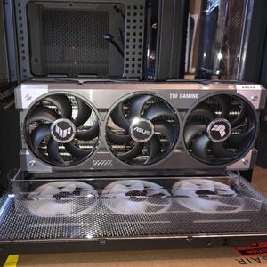 May include: A gray ASUS TUF Gaming graphics card with three black cooling fans installed horizontally inside a computer case. Below the graphics card, three white cooling fans are visible through a clear plastic tray on a black mesh base.