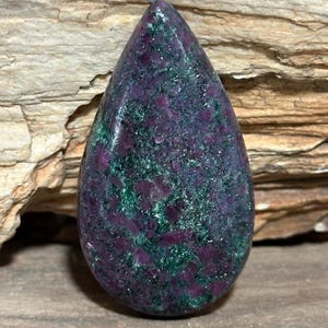 May include: A teardrop-shaped cabochon gemstone featuring a mottled pattern of deep magenta-purple and dark forest green mineral inclusions. The surface is polished to a smooth, reflective finish, showcasing a dense, crystalline texture throughout.