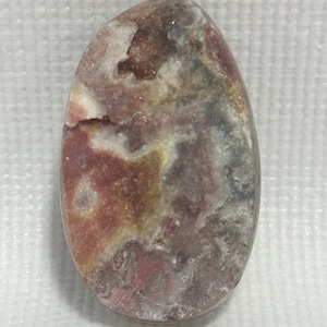 May include: An oval-shaped polished gemstone cabochon featuring a marbled pattern of dusty rose, muted red, cream, and charcoal gray tones. The surface displays a natural crystalline cavity near the top edge with a textured, uneven appearance.