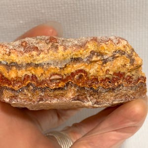 May include: A close-up of a layered rock formation with bands of orange, yellow, and brown. The rock has a rough, textured surface and is held in a hand with a silver ring.