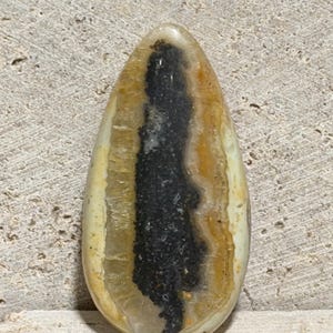 May include: A teardrop-shaped polished gemstone cabochon featuring a prominent, elongated black center core surrounded by concentric bands of translucent amber, cream, and beige mineral layers with a smooth, glossy, reflective surface finish.