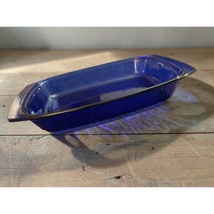 May include: A rectangular, cobalt blue glass baking dish with a textured pattern. The dish has a silver-coloured rim and handles. The dish is empty and sits on a wooden surface.