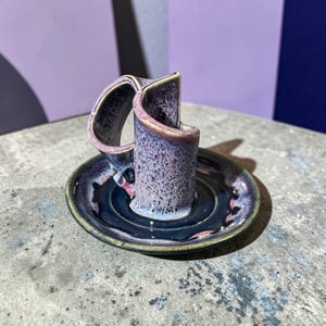 May include: A handcrafted ceramic candle holder with a unique design. The holder features a cylindrical body with a curved handle, all in a speckled purple glaze. It sits on a dark blue saucer with a glossy finish, perfect for home decor.