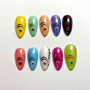 May include: A set of ten almond-shaped artificial nails, each painted with a unique design. The designs feature a central eye motif with curved lines and dots, set against a variety of colors including yellow, green, orange, purple, and pink.
