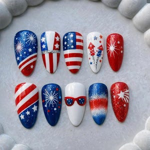 May include: A set of ten artificial nails with patriotic designs. The nails feature red, white, and blue colors with patterns including stars, stripes, fireworks, and the American flag. The nail shapes are oval.