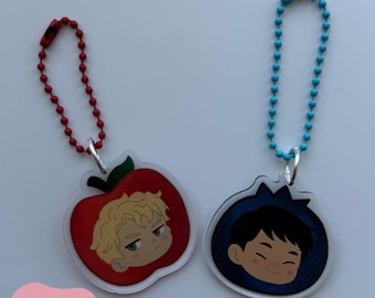 Puede incluir: Dos llaveros de acrílico con el texto FRUIT KEYCHAINS y HEATED RIVALRY. Uno muestra una manzana roja con un personaje rubio, el otro una fruta azul oscuro con un personaje de pelo oscuro. Tienen cadenas de bolas de color rojo y azul.