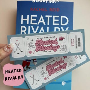 Könnte beinhalten: Ein blaues Buch mit dem Titel "Heated Rivalry" und ein passendes Lesezeichen. Das Lesezeichen ist wie ein Ticket gestaltet und zeigt Hockey-Illustrationen und den Text "Heated Rivalry Book Club."
