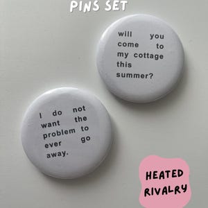 May include: Two white circular pins with black text. One pin says "I do not want the problem to ever go away." The other pin says "will you come to my cottage this summer?" A pink sticker reads "HEATED RIVALRY."