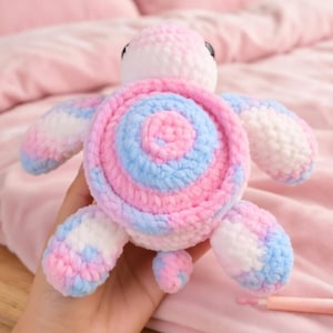Könnte beinhalten: Ein Plüschschildkrötenspielzeug aus weichem Garn in einem bunten Muster aus Pastellrosa, Hellblau und Weiß. Der Panzer hat ein kreisförmiges, gewirbeltes Design, während der Kopf zwei kleine schwarze Augen hat. Die Gliedmaßen und der Schwanz sind abgerundet.