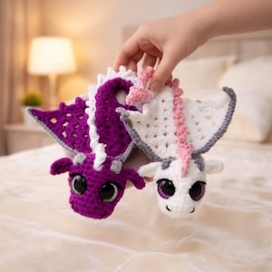 Könnte beinhalten: Zwei gehäkelte Drachen-Stofftiere, eines in Dunkelviolett mit weißem Rückenkamm und eines in Weiß mit rosa Rückenkamm. Beide haben große, dunkle, glänzende Augen, kleine Hörner und breite, strukturierte Flügel. Der violette Drache hat silberne Akzente.