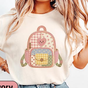 May include: An ivory t-shirt featuring a central graphic of a patchwork-style backpack. The backpack design incorporates various patterns including gingham, polka dots, and floral prints in pastel pink, yellow, blue, and green, with a heart face.