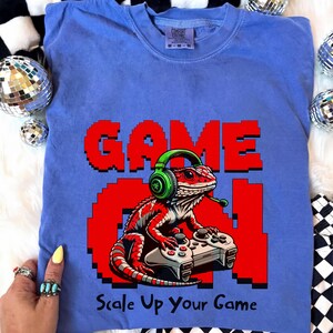 Puede incluir: Camiseta azul con un gráfico rojo y blanco de un lagarto con auriculares y un mando de videojuegos. Las palabras "GAME ON" están en letras pixeladas rojas, con la frase "Scale Up Your Game" debajo.