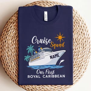Puede incluir: Camiseta azul marino con un diseño de crucero. El diseño incluye las palabras "Cruise Squad", un sol, palmeras y un delfín. También aparece el texto "Our First Royal Caribbean".