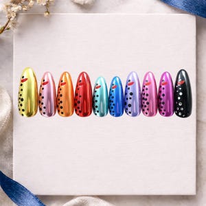 May include: A collection of ten metallic nail tips in various colors, including gold, pink, orange, red, teal, blue, purple, magenta, and black. Each nail features a small red accent and black dot pattern, arranged in a row.