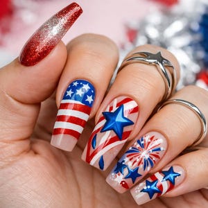May include: Close-up of a hand with long, decorated nails. The nails feature patriotic designs with red, white, and blue colors, including the American flag, stars, and fireworks. One nail has a red glitter gradient.