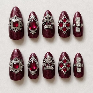 May include: A set of ten burgundy-colored artificial nails with a glittery finish. Each nail features ornate silver-colored embellishments, including square, teardrop, and crown-shaped designs, and red jewel accents.