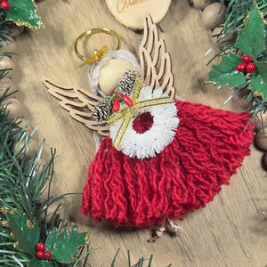 May include: A handmade Christmas angel ornament with a red yarn dress, wooden wings, and a gold halo. The angel has a white yarn collar and is decorated with a small pinecone and red berries. The ornament is part of a Christmas wreath.