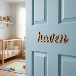 May include: Wooden sign spelling "haven" in a cursive font, fixed to a light blue door. The sign is made of light-coloured wood. The background shows a cot and a colourful rug.