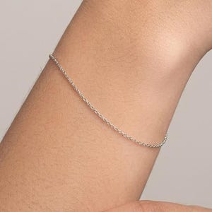 May include: A delicate, thin silver-toned chain bracelet worn around the wrist. The bracelet features a simple, minimalist link design with a polished metallic finish that catches the light, creating a subtle and refined appearance on the skin.