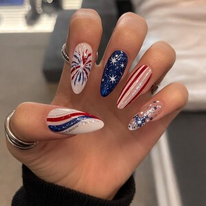 May include: A hand with long, almond-shaped nails, each painted with a patriotic theme. Designs include a firework, a starry night, red and white stripes, and a silver glitter accent. The color palette features red, white, and blue.