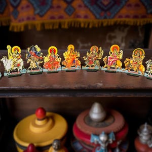 May include: A collection of colorful, illustrated figurines depicting Hindu deities. Each figure has a golden halo and is mounted on a small wooden base. The figurines are arranged on a dark wooden shelf, with other decorative items visible in the background.