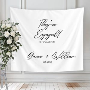 May include: White fabric backdrop with the text "They're Engaged!" in elegant script. Below, it reads "Let's Celebrate!" and the names "Grace + William" with an established date. A floral arrangement is on the left.